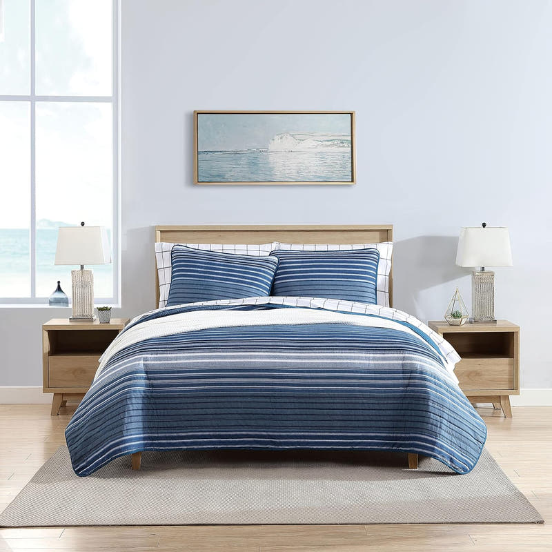 A 3-piece cotton-blend quilt set with a reversible design, featuring a striped pattern, displayed on a bed in a room with a seascape painting on the wall and a wooden bedframe.